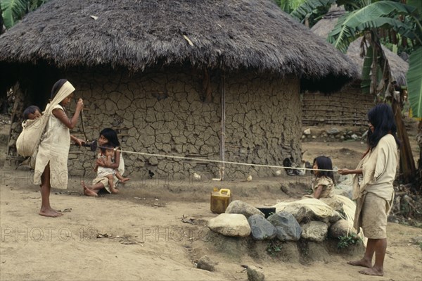 COLOMBIA, Sierra Nevada de Santa Marta, Avinque, Parque Nacional. Kogi-Wiwa family spinning rope from strands of fique cactus fibre. Mother holds spindle and spins whilst father holds out extended new rope. Two young daughters look on whilst baby son sleeps in large woven cotton mochila/shoulder bag on mothers back  his weight taken by broad headband. All family wearing traditional woven cotton mantas/cloaks