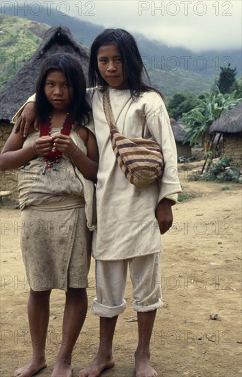 COLOMBIA, Sierra Nevada de Santa Marta, Sierra Avingue, Parque Nacional. Portrait of Kogi brother and sister standing in village of Avingue with grass thatched huts behind them Wearing traditional woven cotton garments Boy has typical fique/cactus-fibre mochila/shoulder bag with brown bands from natural root dye