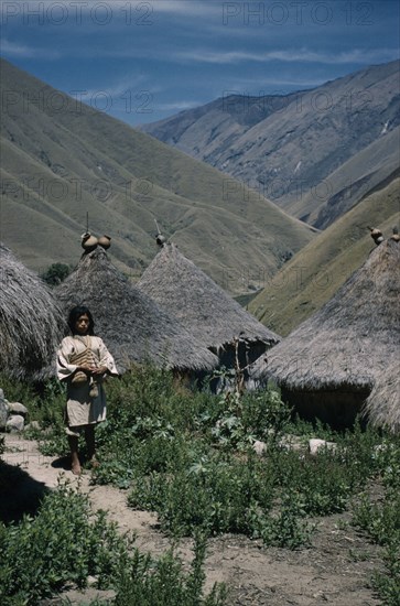 COLOMBIA, Sierra Nevada de Santa Marta, Kogi Tribe, "San Miguel village. A young “mama” / priest stands in pathway leading  to thatched dwellings and ritual centres, with sacred pots at roof apices."