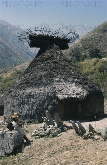 COLOMBIA, Sierra Nevada de Santa Marta, Kogi Tribe, A “nuhue” / temple at ritual centre of Macotama. Granite seats placed outside temple for “mamas” / priests. Potsherds in the rack at roof apex. Young Kogi “vasayo”  / commoner leans on granite boulder