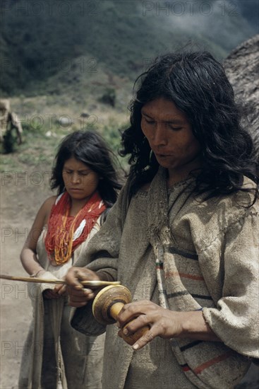 COLOMBIA, Sierra Nevada de Santa Marta, Kogi Tribe, "Man rubs lime encrusted gourd with ritual stick, having inserted lime  powder into a wad of coca leaves held in his cheek. Process has a ritual/sexual significance – the stick is the male penis, the gourd the female vagina. His wife stands behind wearing strings of red and yellow glass beads – a sign of wealth"