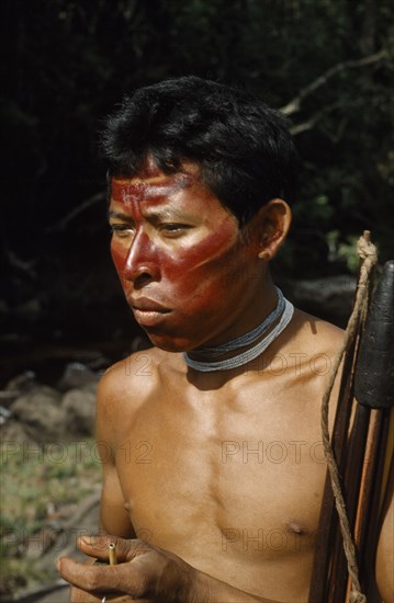 COLOMBIA, Vaupes Region, Tukano Tribe, "Hunter with red “karajuru” / facial paint, wooden ear plug and white glass bead necklace, carrying coca powder pouch with bone pipe – coca sustains whilst hunting. Hard “macana” wood bow and quiver of curare-tipped arrows in resin-coated sheath to protect from rain."