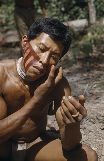 COLOMBIA, Vaupes Region, Tukano Tribe, "Man applies red “karajuru” facial paint, wearing precious strings of white glass beads and a bone ring, with coca pouch slung over shoulder."