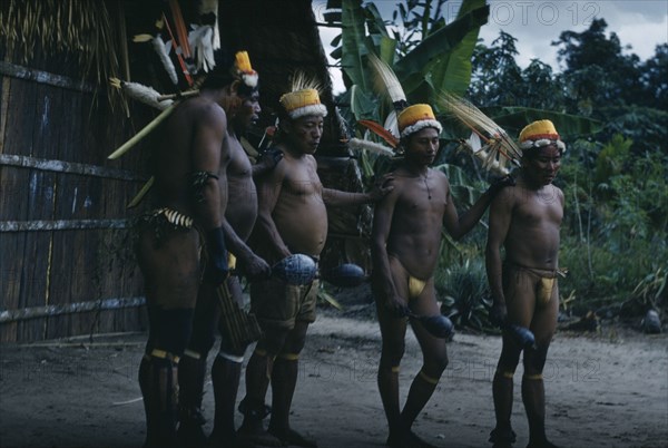 COLOMBIA, Vaupes Region, Tukano Tribe, "Men perform the Maraca Dance outside “maloca” wearing feather head-dresses, bead nut amulets, festive facial and body paint,woven fibre garters, nut ankle rattles, monkey tooth belts, green sweet-smelling herbs and waving finely engraved maracas."
