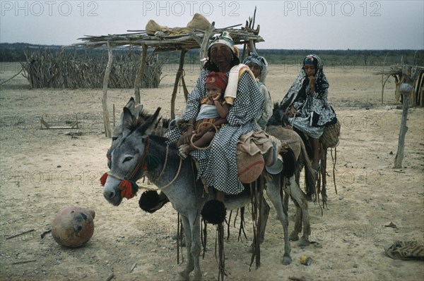 COLOMBIA, Guajira Peninsula, Guajiro / Wayuu Tribe, "Mother and daughters on donkeys, normal means of transport in Guajira. Mother protects face from strong sun and salty winds with mixture of burnt fungi and goat fat. Note fired clay water vessel on ground and cactus-wood goat enclosure in background"