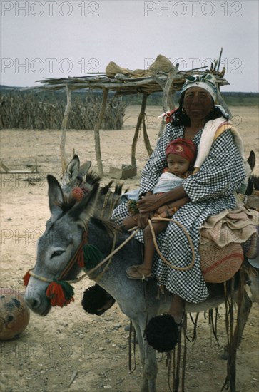 COLOMBIA, Guajira Peninsula, Guajiro / Wayuu Tribe, "Portrait of Wayuu mother with baby daughter. Facial protection against strong sun and wind from mixture of burnt fungi and goat fat. Mother wears woollen pom-poms on feet to protect toes and as adornment, donkey also decorated with woollen pom-poms."