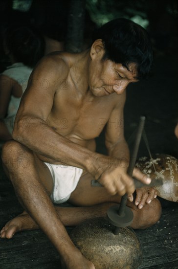 COLOMBIA, Choco Region, Noanama Tribe, Man drills holes in a gourd to make a colander for straining fruit to be made into “chicha” beer. Behind his leg another gourd is engraved with ritual figures
