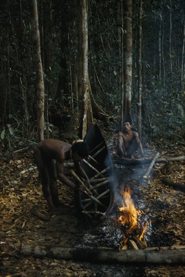 COLOMBIA, Vaupes Region, Tukano Tribe, "2 men making a canoe from a single tree trunk. After initial shaping with axes, canoe burnt and sides expanded in heat with wooden spacers. Burning slims sides, shapes and hardens the wood"