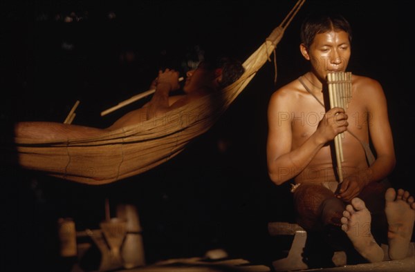 COLOMBIA, Vaupes Region, Tukano Tribe, "Boys playing panpipes at dusk in entrance to maloca / longhouse. One sits on an elders stool, other lies in a “cumare” / fibre hammock"