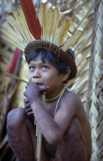 COLOMBIA, Vaupes Region, Tukano Tribe, Young boy playing panpipes outside maloca prior to a festival. Painted with deep purple we leaf dye and wearing crown of red macaw and yellow toucan feathers