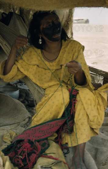 COLOMBIA, Guajira Peninsula, Guajiro / Wayuu Tribe, "Woman embroiders a “faja” / woven woollen belt for her husband, sitting on a “fique” / cactus fibre hammock; face smeared with black fungal paint mixed with goat fat."