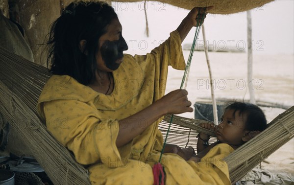 COLOMBIA, Guajira Peninsula, Guajiro / Wayuu Tribe, Mother and baby son in a “fique” / cactus fibre hammock in their desert home. Mother with black fungal facial paint to protect against strong wind and sun. She sews a “faja” / woven woollen belt for husband. Baby wears amulet with charms.