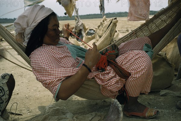 COLOMBIA, Guajira Peninsula, Guajiro / Wayuu Tribe, Wayuu woman sews a coloured “faja” / woven woollen belt for her husband sitting in a fique / cactus fibre hammock in their scanty desert shelter