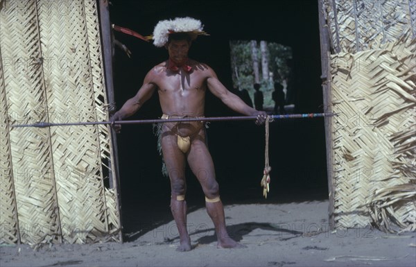 COLOMBIA, Vaupes Region, Tukano Tribe, "Shaman in full festive regalia – feather head-dress, red “karajuru” facial paint, dark purple “we” body dye, monkey tooth belt, “cumare”  fibre garters painted with yellow clay. He beats and waves the “kurubeti” pebble stave to ward off evil spirits and thunder."