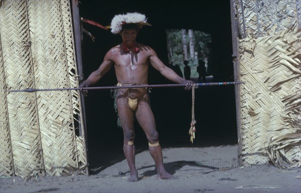 COLOMBIA, Vaupes Region, Tukano Tribe, "Shaman in full festive regalia – feather head-dress, red “karajuru” facial paint, dark purple “we” body dye, monkey tooth belt, “cumare” fibre garters painted with yellow clay. He beats and waves the “kurubeti” pebble stave to ward off evil spirits and thunder."