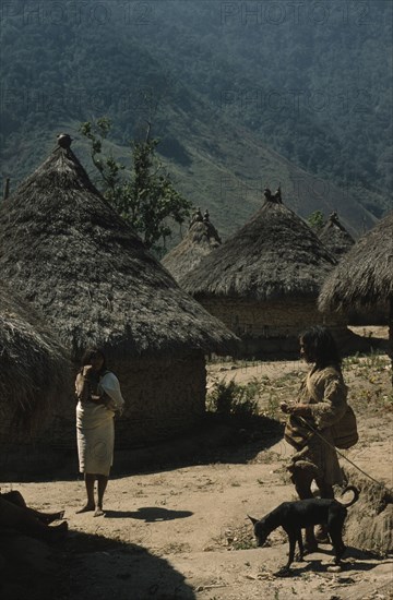 COLOMBIA, Sierra Nevada de Santa Marta, Kogi Tribe, "San Miguel village, important Kogi centre, thatched roofs with pots of apex.  Man with dog wearing threadbare woven “manta” / cloak and two “mochilas” / shoulder bags, talking to woman with new “manta” and red bead necklace"