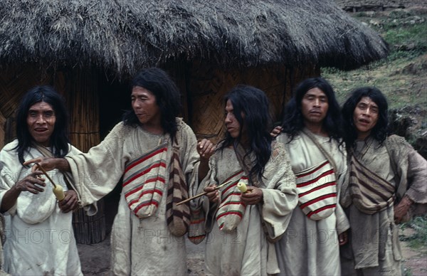 COLOMBIA, Indigenous Tribes, Kogi, Chendukua. A group of Kogi men outside a grass-thatched village home on S.side of Sierra. Drunk on aguadiente/distilled sugarcane liquor -sold to Indians by Colombian campesinos/peasants Hand-woven white cotton and darker fique/cactus-fibre mochilas/bags around necks. Two carrying lime gourds and sticks to transfer powdered lime into wads of coca-leaves in their mouths - lime acts as catalyst to release very small amount of cocaine alkaloid.