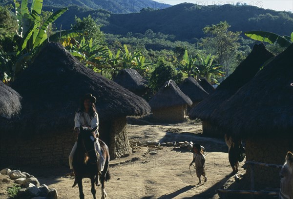 COLOMBIA, Sierra Nevada de Santa Marta, Avingue, "Parque Nacional. Kogi Wiwa leader riding his horse through Avingue village between mud-wattle walled and grass thatched homes with children playing,  banana palms and cultivation plots surrounding village.  "