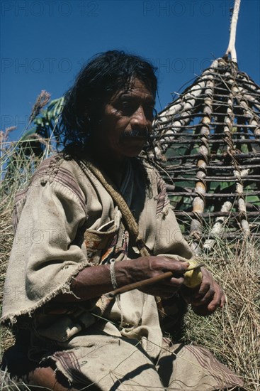 COLOMBIA, Sierra Nevada de Santa Marta, San Antonio, "Mama/Priest Valencia takes a ""coca break"" - taking lime powder from his poporo/gourd to put into wad of coca leaves in his mouth - whilst thatching the roof of his house Wearing traditional thick woven cotton manta/cloak Indigenous Tribes American northern Caribbean slopes of Sierra  "