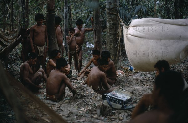 COLOMBIA, Llanos Plains, Agua Clara, "Cuiva: First Contact with Agua Clara  group of men listening to tape recording of their Cuiva ""cousins""  a group contacted 2 years before in 1968 but now cut off by Llanero/cowboy cattle lands.  French Canadian anthropologist Bernard Arcand with hammock and mosquito net strung above him Cuiva cumare/ fibre hammocks in trees behind At this stage Cuiva had no western clothing  just bark cloth guayukos/aprons   Llanos Plains running East from Andes to Orinoco. rio Agua Clara Indigenous Tribes Cuiva   "