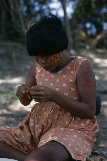 COLOMBIA, Llanos Plains, Rio Meta Arauca, "rio Meta ""Tree Camp"". Cuiva woman  Yanakwa's wife  wearing traded flower print dress with red Achiote facial paint She threads coloured glass beads obtained from boats which ply the rivers of the Llanos  buying local produce and selling trade goods  Llanos plains running East from Andes to Orinoco. rio Meta  Arauca Indigenous Tribes Cuiva  "