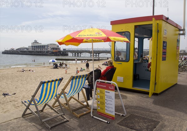 ENGLAND, Dorset, Bournemouth, Lifegaurd station on The East Beach with Bournemouth Pier beyond tourists on the sandy beach and paddling in the sea.