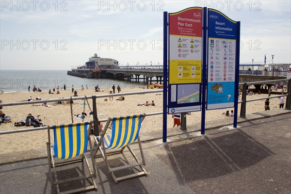 ENGLAND, Dorset, Bournemouth, Deckchairs beside an information sign on The East Beach promenade with Bournemouth Pier beyond tourists on the sandy beach and paddling in the sea.