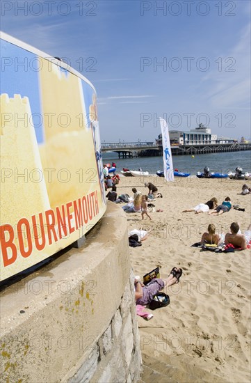 ENGLAND, Dorset, Bournemouth, Tourists on the sand of the West Beach beside the pier. A sign reading Bournemouth is in the foreground