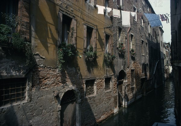 ITALY, Veneto, Venice, "Canalside houses with crumbling plaster and brickwork, plants and flowers in window boxes and washing hanging between buildings overhead."