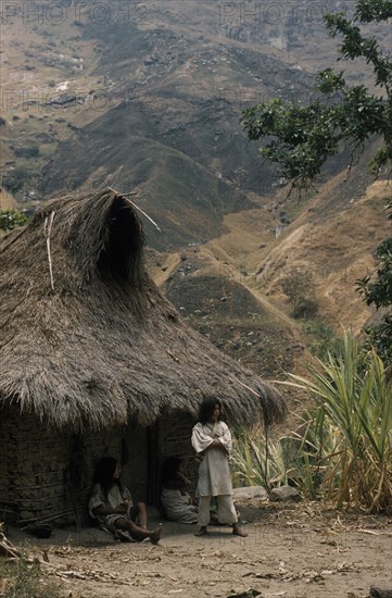 COLOMBIA, Sierra Nevada de Santa Marta, Kogi Tribe, "Young man stands outside the family home above Takina, wearing typical woven cotton manta / cloak and trousers with crossed “mochilas”  / shoulder bags of “fique” cactus fibre."