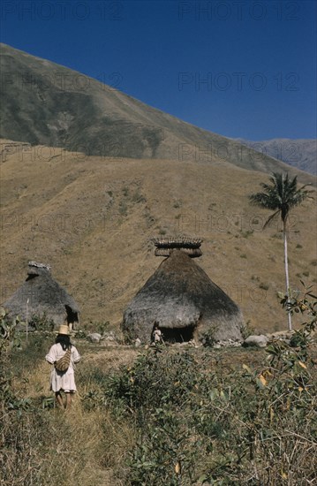 COLOMBIA, Sierra Nevada de Santa Marta, Kogi Tribe, "Religious centre of Takina, high in Sierra Nevada. Man wearing hand woven and hand dyed “mochila” / shoulder bag, walks to meet a “mama” / priest emerging from the “nuhue” / temple, with large rack of potsherds at the roof apex"