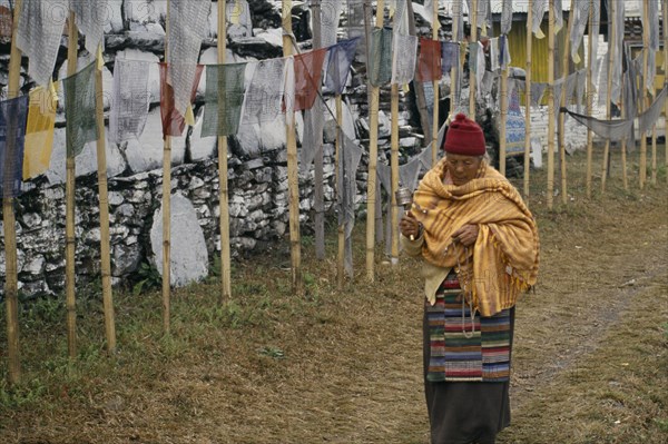 INDIA, Sikkim, Tashiding, "A pilgrim holding a prayer wheel and beads, walking past a mani wall and Buddhist prayer flags."