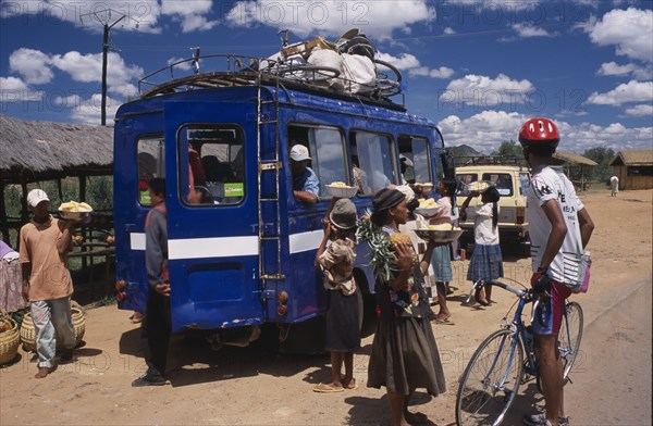 MADAGASCAR, Transport, Road to Antsirabe. Minibus at a roadside stopping point with a woman and children selling pineapple from bowls to the passengers onboard and a man wearing a helmet  ona bicycle in the foreground.