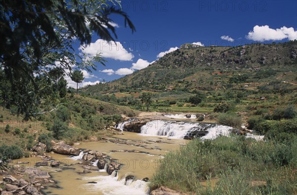 MADAGASCAR, Landscape, Road to Antsirabe. River and waterfall running between green landscape with buildings set into the hillside