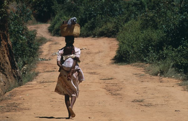 MADAGASCAR, People, Women, Near Ambositra. Portrait of a woman carrying a baby in a sling on her back and wicker baskets on her head walking up a red earth road