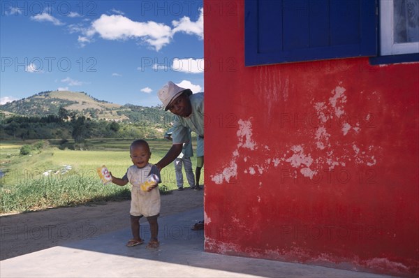 MADAGASCAR, People, Road to Fianarantsoa. Toddler with Grandmother appearing from around exterior wall of a roadside cafe with Classiko red and blue livery and lush green landscape and hills in the distance behind them