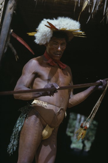 COLOMBIA, Vaupes Region, Tukano Tribe, "Shaman beats ceremonial “kurubeti” / stave to fend off evil spirits and thunder storms. Adorned with white egret down and macaw tail feather crown, monkey tooth belt red “karajuru” facial paint and black leaf body dye. Sweet-smelling herbs dangle from his belt"
