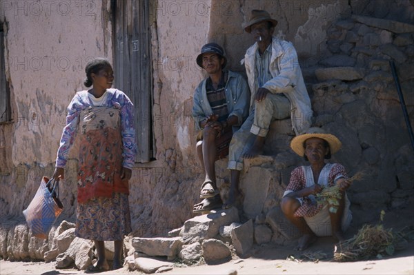 MADAGASCAR, People, Road to Ranomanfana. Two men sitting on steps at side of building next to two women wearing colourful clothing one selling vegetables