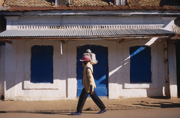 MADAGASCAR, Ambalavao, Boy wearing a red cap carrying a bag on top of his shoulder walking past a white washed building with a blue door and shutters