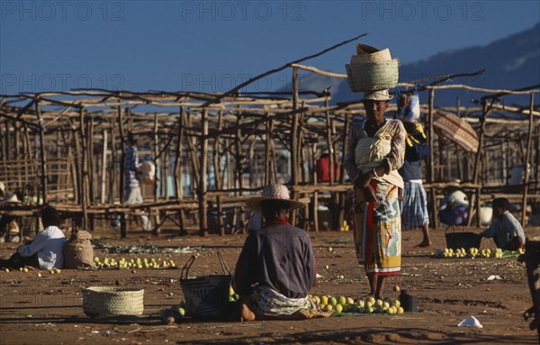 MADAGASCAR, Ambalavao, Two women selling fruit at a sunday market place with empty stalls one woman sat on ground with her back turned and one woman standing carrying wicker baskets on her head facing lens