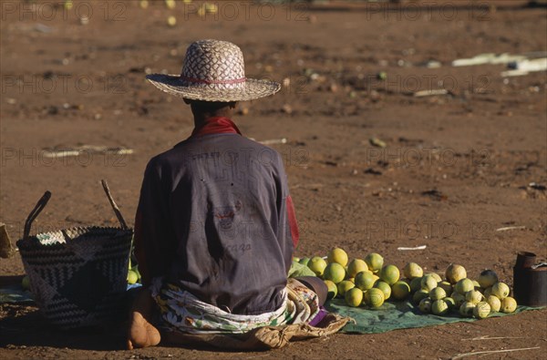 MADAGASCAR, Ambalavao, Woman sat on ground with her back facing the lens wearing straw hat and Chicago Bulls basket ball shirt selling fruit