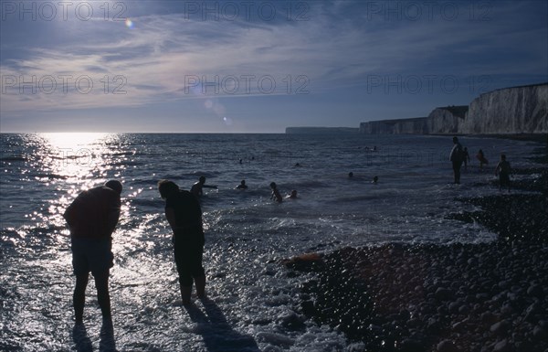 ENGLAND, East Sussex, Birling Gap, People in silhouette paddling at the waters edge on pebbled beach
