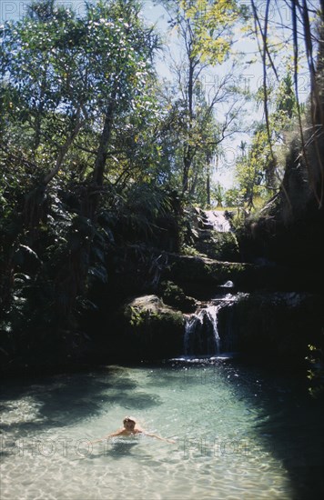 MADAGASCAR, Isalo National Park, Trees surrounding a waterfall running over rocks into a natural fresh pool with a woman swimming in the clear water