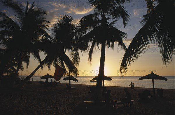 MADAGASCAR, Tulear, Ifaty Beach at sunset with view through palm trees and straw sun shades towards the sea with people on the sand in silhouette
