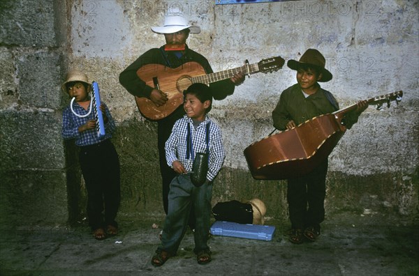 MEXICO, Oaxaca State, Oaxaca, " In a backstreet of the colonial town of Oaxaca stands a local family playing traditional Mexican music to an eclectic group of enthusiastic passer-bys. The father seems to be leading the musical band of four and stands proudly at the back against a grey brick wall. He sports a white cowboy hat and blows on a harpsichord whilst strumming his acoustic guitar. On his right hand side stands his oldest son, dressed in similar attire strumming a much larger guitar that looks more like a cello. On the left of shot, there is the middle son who unenthusiastically leans in the corner, again dressed in a beige cowboy hat, a checkered shirt and dark torusers. He is playing a plastic wind instrument which also appears to have some kind of keyboard. In front of these three stands the youngest of the group. The young child wears a checkered blue and white shirt and seems to be shputing and gesturing at the passing trade for tips or 'propinas'. The ground below them lie...