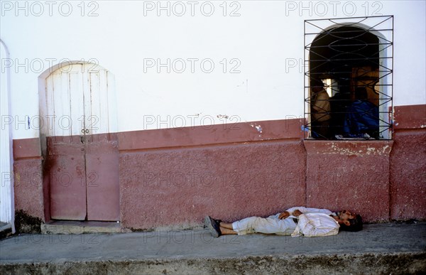 EL SALVADOR, Suchitoto, "In the heart of this old colonial town lies a colourful backstreet. The outside walls of the houses slopes down from right to left and at the front of shot one can make out the edge of a bumpy pavement. The wall in the background has been painted halfway in a rustic red colour. The wooden doorway is visibly closed and there are iron bars in front of the only visable window. Behind the bars is a local barbers as one can just about make out the shapes of a barber and his client sitting down. On the pavement on the foreground lies a local dressed from head to toe in beige linen. He is sleeping on the dusty ground with his legs crossed and his hands lying gently on his chest. His skin is dark and worn and although his position seems a little unorthodox, on the whole he seems surprisingly comfortable. "