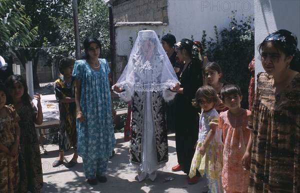 TAJIKISTAN, Wedding, "The bride in a white dress, veil and decorated jacket surrounded by guests at a village wedding."