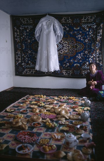 TAJIKISTAN, Wedding, "A white wedding dress hangs on the wall in front of a rug, and a large display of food is laid out awaiting guests from a village wedding"