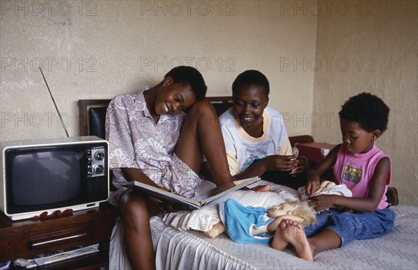 SOUTH AFRICA, Gauteng, Alexandra Township, "Three girls relaxing in bedroom of home.  Two eldest looking at album while youngest plays with white, blonde haired doll. "