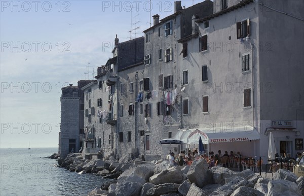 CROATIA, Istria, Rovinj, Old city waterfront building with a washing line hanging from shuttered windows and people sitting outside under the sun awning of a Pizzeria restaurant attached to the bottom of the building. Large sea defence boulders lining the waters edge
